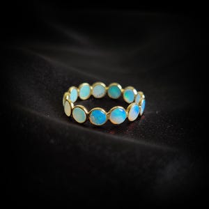 May include: A gold band ring with a row of round, light blue opals. The ring is set on a black background.