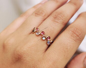 May include: A gold ring with a row of colorful gemstones. The gemstones are pink, white, and gray. The ring is on a person's finger.