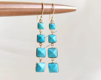 May include: A pair of gold-toned drop earrings featuring three square, light blue turquoise stones. The earrings have a simple hook design and are displayed against a neutral background. The turquoise stones are faceted, adding a touch of sparkle.