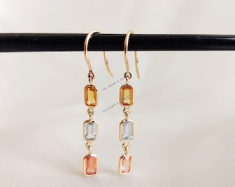 May include: Gold-toned drop earrings featuring three rectangular gemstones in shades of yellow, light blue, and peach. The earrings are suspended from a curved hook and are displayed against a neutral background.
