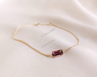 Könnte beinhalten: Ein zartes Armband aus einer goldenen Kette mit einem einzigen rechteckigen rosafarbenen Edelstein. Das Armband liegt auf einem weißen Hintergrund.