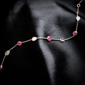 May include: A gold chain necklace with alternating oval-shaped white and pink gemstones. The necklace is on a black fabric background.