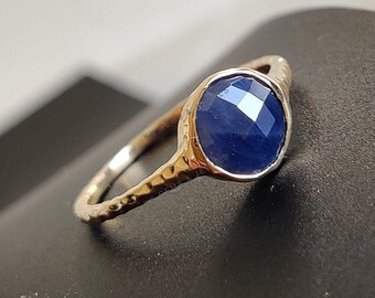 May include: A gold ring with a textured band and a dark blue gemstone. The gemstone is faceted and has a round shape.