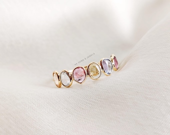 May include: A gold ring with a row of seven oval-shaped gemstones. The gemstones are in a rainbow of colors, including pink, yellow, blue, and clear.