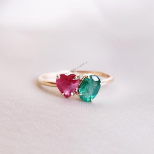 May include: A gold ring with a heart-shaped ruby and a round emerald gemstone. The ring is on a white background.