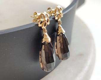 May include: A pair of gold earrings with a teardrop-shaped smoky quartz gemstone dangling from each earring. The earrings have a delicate floral design.
