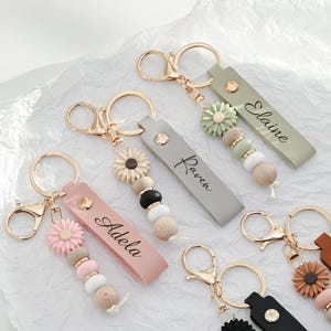 May include: A collection of personalized keychains with gold-colored hardware. Each keychain features a leather strap with a name, a decorative flower, and a series of wooden and colored beads. The keychains are in various colors, including pink, green, and brown.
