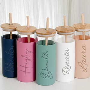 Puede incluir: Cinco vasos de vidrio personalizados con tapas y pajitas de bambú. Cada vaso tiene una funda de color diferente: azul marino, rosa, verde azulado, blanco y melocotón. Cada vaso está inscrito con un nombre diferente en cursiva: Shirley, Hayley, Demilla, Renata y Laura.