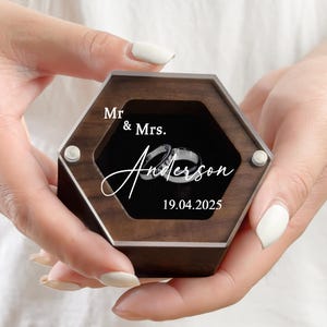 Könnte beinhalten: Eine sechseckige Holz-Ringbox mit einem transparenten Acryldeckel, die zwei silberne Ringe enthält. Die Box ist mit "Mr. & Mrs. Anderson 19.04.2025" graviert. Die Box wird in den Händen einer Person gehalten.