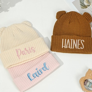 May include: Three knitted beanies with bear ears. One is cream with "Paris" embroidered in pink, another is pink with "Laird" in blue, and the third is brown with "HAINES" in white. A wooden owl toy is also present.
