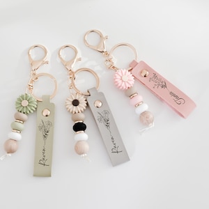 Op de afbeelding: Vier sleutelhangers met goudkleurige hardware. Elke sleutelhanger heeft een ander kleurenschema, een leren label met bloemenillustraties en het woord "Raven". Elke sleutelhanger heeft een bloemenbedel en houten kralen.