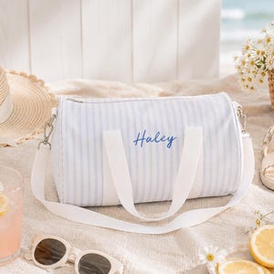 May include: A light blue and white striped duffel bag with the name "Haley" in blue script. The bag has white straps and a white shoulder strap. The scene includes a straw hat, sunglasses, a drink, sandals, and flowers.