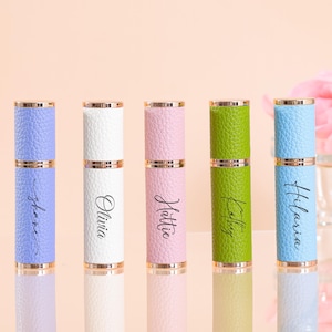 May include: Five personalised perfume atomisers in lavender, white, pink, green, and light blue. Each has a gold top and bottom, with a name in cursive script. The atomisers are for perfume or fragrance.