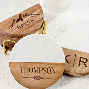 Pode incluir: Conjunto de bases personalizadas com um design branco e de madeira. As bases apresentam nomes como "Brian" e "Thompson" com desenhos de montanhas e geométricos. São exibidas em um suporte de metal dourado.
