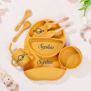 May include: Mustard yellow baby feeding set with the name "Sophia" printed on the plate and bib. The set includes a divided plate, bowl, cup, bib, and utensils. A planet graphic is on the plate and cup.