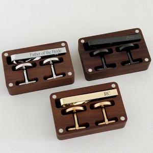 Op de afbeelding: Drie sets manchetknopen en stropdasklemmen in houten dozen. Eén set is zilverkleurig met de gravure "Father of the Bride". Een andere is zwart met een handtekening. De derde is goudkleurig met "RC".
