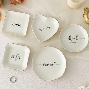 May include: A selection of white ceramic ring dishes in various shapes: square, heart, and round. Each dish has a gold rim and personalised text in black, including initials, names, and a date. A silver ring is also present.