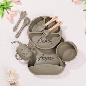 May include: A set of baby feeding essentials in a muted green color. The set includes a divided plate, bowl, bib, cup, and utensils. The name "Aaron" is printed on the plate, bib, and utensils. An elephant graphic is on the cup and plate.