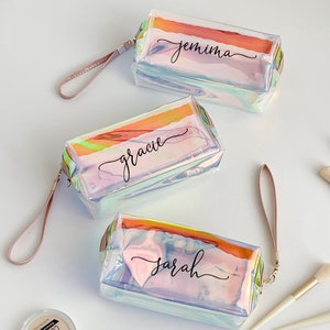 Può includere: Tre trousse per cosmetici iridescenti con i nomi "Jemima", "Gracie" e "Sarah" in corsivo nero. Ogni trousse ha un cinturino da polso abbinato e una forma rettangolare. Sono su una superficie bianca.