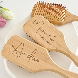 May include: Three wooden hairbrushes with light brown bristles and handles. Two brushes are engraved with names and the word "Bridesmaid". The brushes are on a white surface, with flowers in the background.