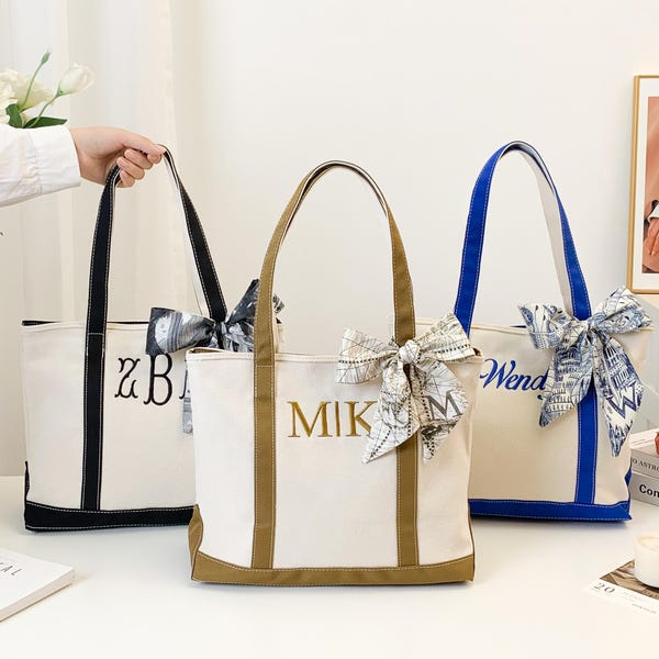 Op de afbeelding: Drie canvas draagtassen met gepersonaliseerde borduursels en decoratieve strikken. De tassen hebben zwarte, bruine en blauwe accenten, met geborduurde namen of initialen. De tassen staan op een wit oppervlak.