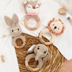 Può includere: Una collezione di sonagli per bambini fatti a mano all'uncinetto a forma di cervo, coniglio, leone ed elefante. Ogni sonaglio ha un anello di legno ed è personalizzato con un nome. Il cervo ha una sciarpa rosa e il leone ha una criniera beige.