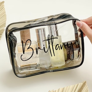 Op de afbeelding: Een transparante, rechthoekige make-uptas met een zwarte ritssluiting en rand, gevuld met diverse parfumflesjes. De naam "Brittany" staat in zwart script over de tas geschreven. De tas wordt door een hand vastgehouden.