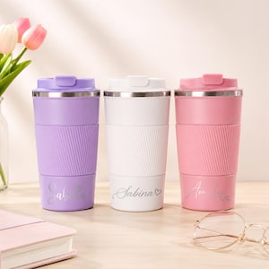 May include: Three personalised travel mugs in lavender, white, and pink, each with a silver lid and a ribbed silicone band. The mugs have names engraved on them. A bouquet of tulips and a notebook are in the background.