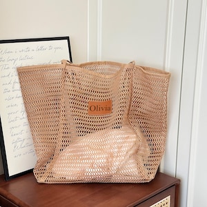 May include: A large, open-weave tote bag in a light brown hue. The bag features a rectangular leather patch with the name "Olivia" on it. A framed print with handwritten text is visible in the background.