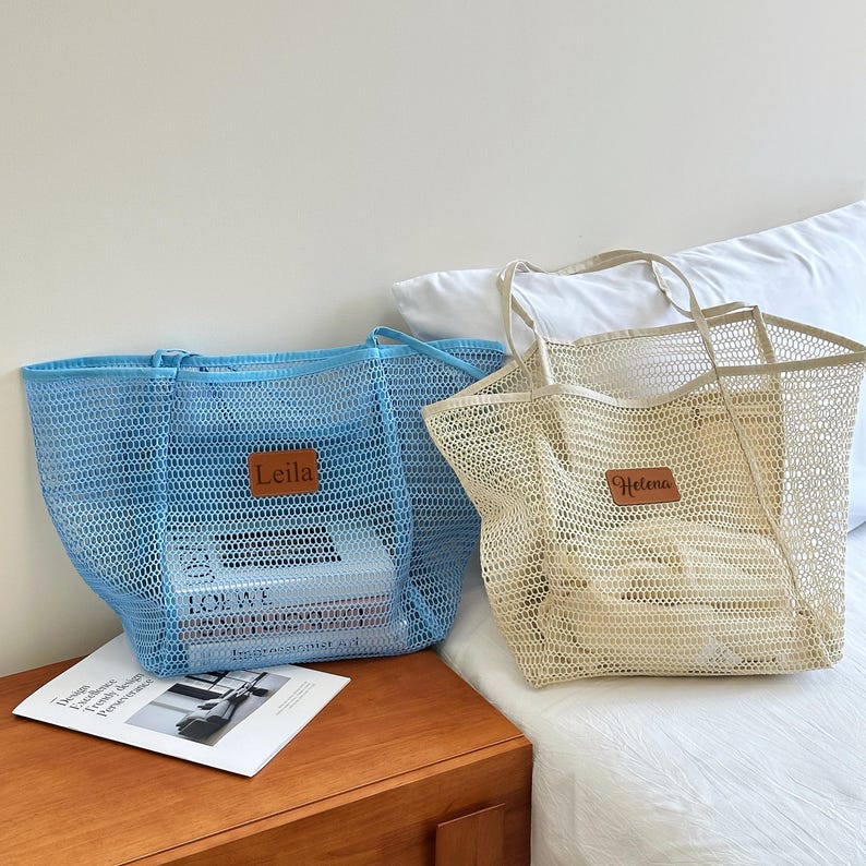 May include: Two mesh tote bags, one light blue and one beige, sit side-by-side. Each bag has a rectangular leather patch with a name on it. The blue bag contains books. The bags are on a bed with a wooden nightstand.