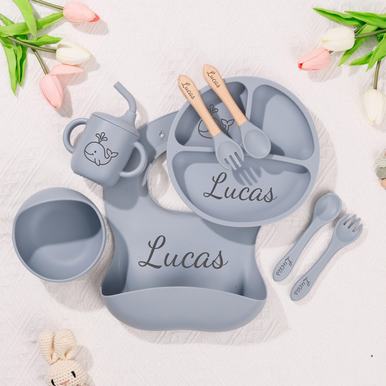 May include: A baby feeding set in light blue, including a divided plate, bowl, bib, sippy cup, and utensils. The set is personalized with the name "Lucas". The utensils have wooden handles. A whale illustration is on the cup.