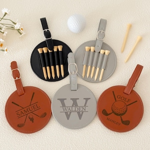 Op de afbeelding: Een verzameling gepersonaliseerde golftaslabels in bruin, zwart en grijs. Elk label heeft een houder voor golftees en een leren riem. Sommige labels zijn gegraveerd met namen of het woord "GOLF".