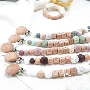 Puede incluir: Primer plano de varios clips de chupete personalizados para bebés. Cada clip presenta cuentas de madera que deletrean nombres como "KATE" y "EMILY", con una variedad de cuentas de colores pastel y neutros. Los clips tienen cierres de madera y están sobre un fondo blanco.