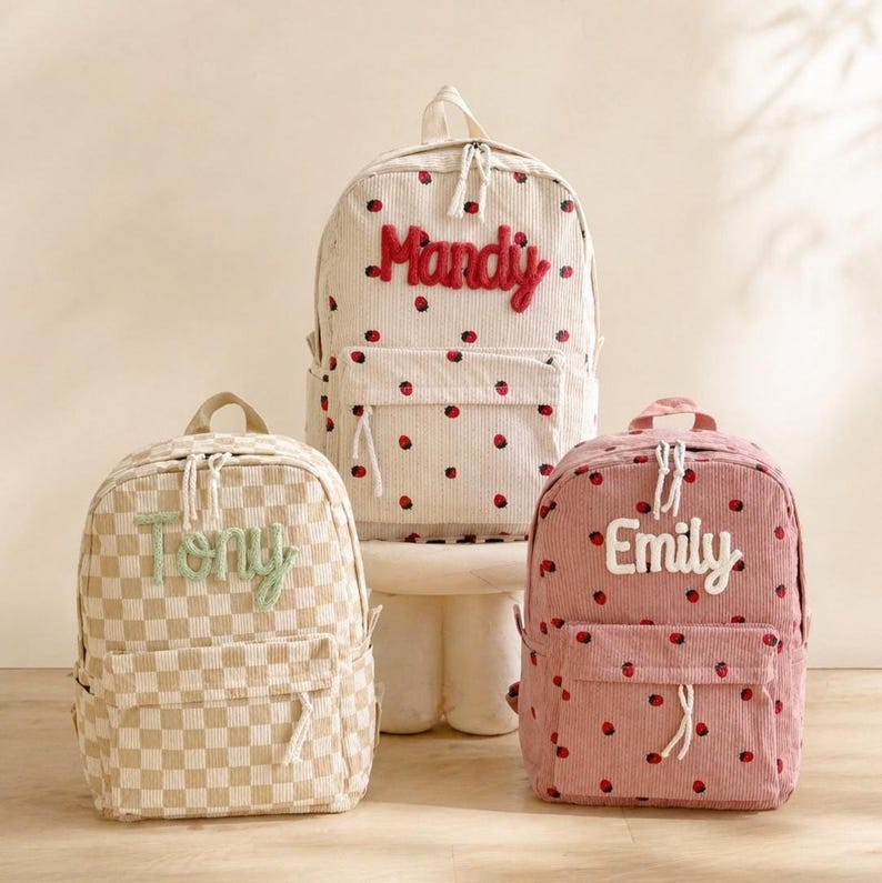 Puede incluir: Tres mochilas personalizadas. Una es beige a cuadros con "Tony" en verde. Otra es blanca con lunares rojos y "Mandy" en rojo. La tercera es rosa con lunares rojos y "Emily" en blanco.