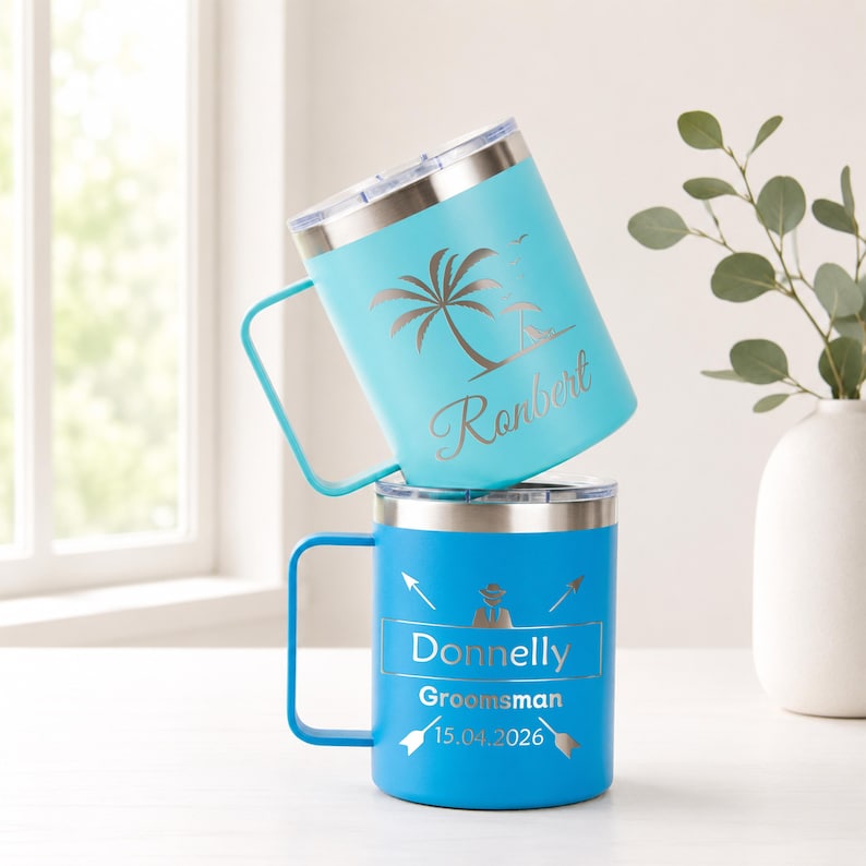 May include: Two insulated travel mugs. One is light blue with a palm tree and the name "Ronbert". The other is blue with the name "Donnelly", the word "Groomsman", and the date "15.04.2026". Both mugs have silver rims and handles.
