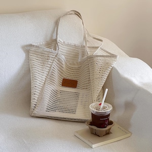 May include: A beige mesh tote bag with a brown leather patch that reads "Kaley". The bag has two handles and is sitting on a white couch. A cup of iced coffee and a magazine are next to the bag.