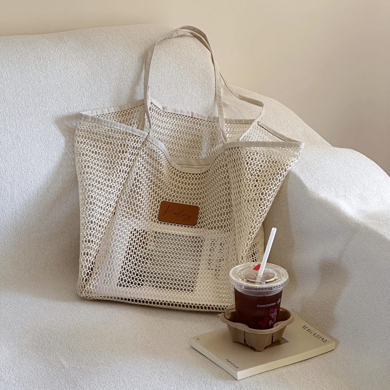 May include: A beige mesh tote bag with a brown leather patch that reads "Kaley". The bag has two handles and is sitting on a white couch. A cup of iced coffee and a magazine are next to the bag.
