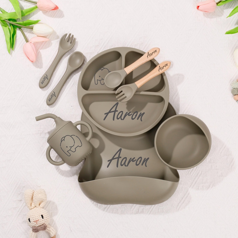 May include: A set of baby feeding essentials in a muted green color. The set includes a divided plate, bowl, bib, cup, and utensils. The name "Aaron" is printed on the plate, bib, and utensils. An elephant graphic is on the cup and plate.