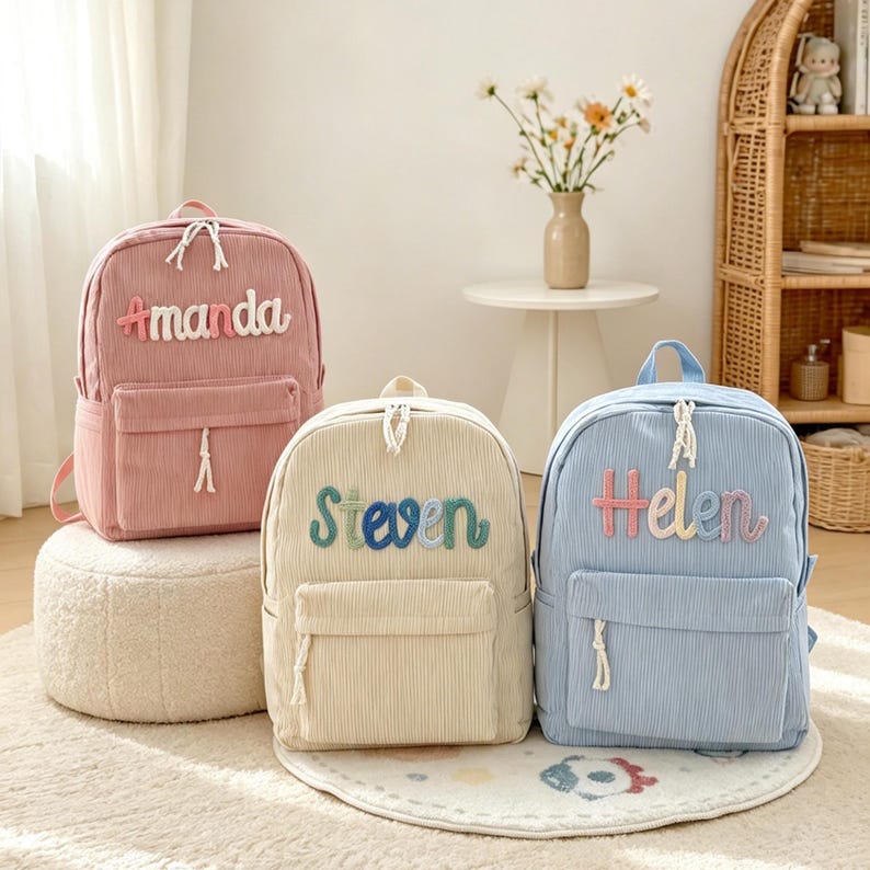Puede incluir: Tres mochilas de pana en rosa, crema y azul. Cada mochila tiene un bolsillo frontal y los nombres "Amanda", "Steven" y "Helen" bordados con hilo colorido. Las mochilas est&aacute;n sobre una alfombra y un peque&ntilde;o puf.