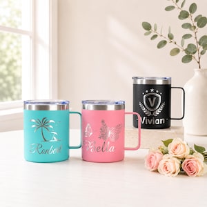 May include: Three stainless steel travel mugs with silver rims and handles. One is teal with a palm tree design and the name "Ronbert". Another is pink with a butterfly design and the name "Bella". The third is black with a shield design and the name "Vivian".