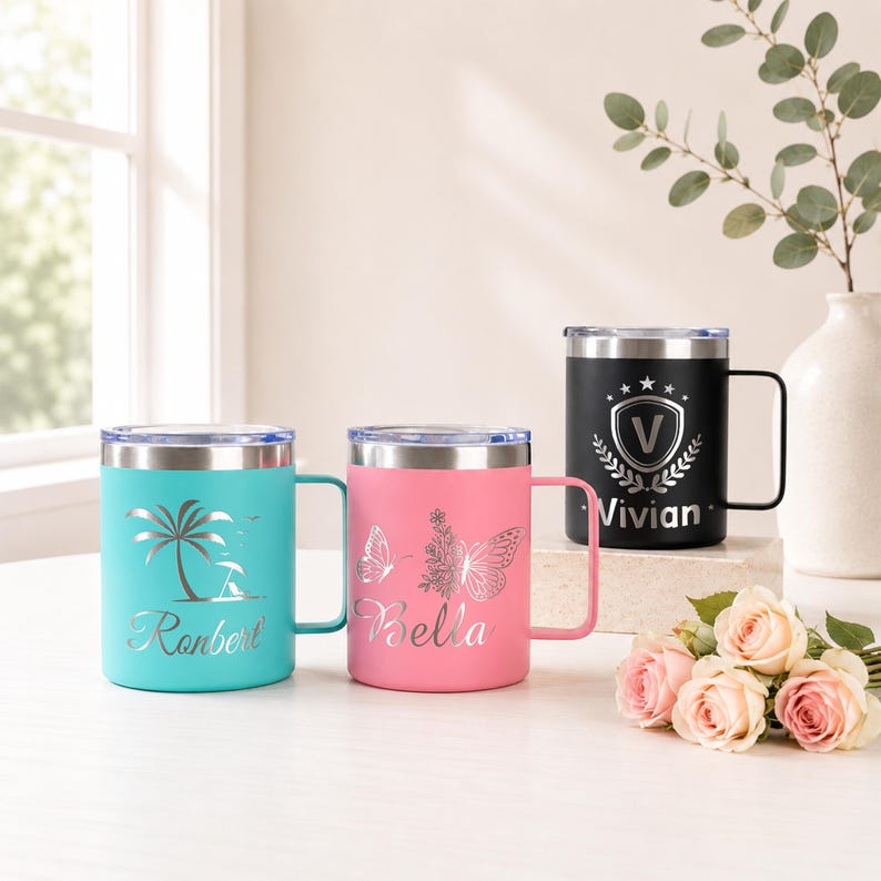 May include: Three stainless steel travel mugs with silver rims and handles. One is teal with a palm tree design and the name "Ronbert". Another is pink with a butterfly design and the name "Bella". The third is black with a shield design and the name "Vivian".