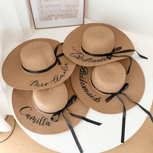 Könnte beinhalten: Vier beige Strohhüte mit breiten Krempen und schwarzen Bändern. Jeder Hut hat einen individuellen Text in Schwarz gestickt: "Mrs Amelia", "Belinda", "Bridesmaids" und "Camilla". Die Hüte sind auf einer weißen Oberfläche angeordnet.