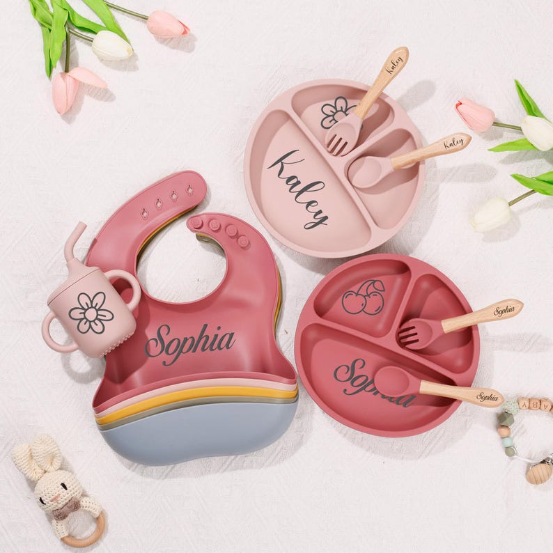 May include: A set of baby feeding essentials in pink and red hues. Includes a bib, divided plates, cups, and wooden utensils. The bib and plates are personalized with names. The utensils have the names engraved on them.