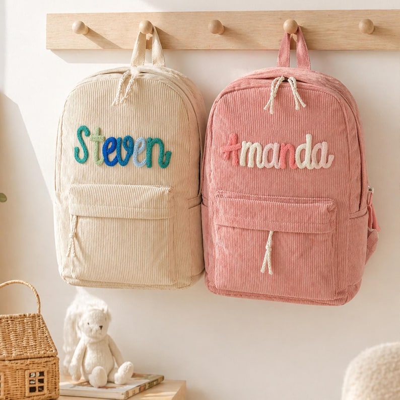 Puede incluir: Dos mochilas de pana, una beige y otra rosa, cuelgan de un perchero de madera. La mochila beige tiene el nombre "Steven" en letras azules y verdes. La mochila rosa tiene el nombre "Amanda" en letras blancas y rosas.
