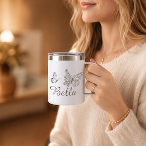 May include: White travel mug with a silver rim and handle. The mug features a butterfly and floral design with the name "Bella" in script. The mug is designed to keep beverages hot or cold.
