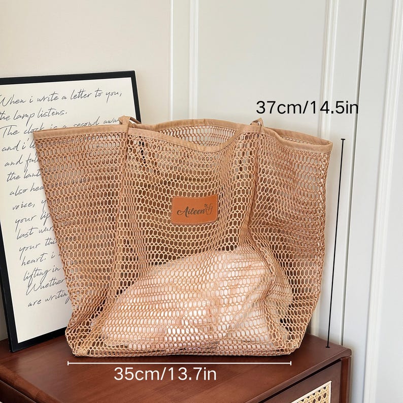 May include: A beige mesh tote bag, 14.5 inches tall and 13.7 inches wide, with a leather patch that reads "Aileen". The bag is filled with a light-colored item. The text "When I write a letter to you..." is visible in the background.