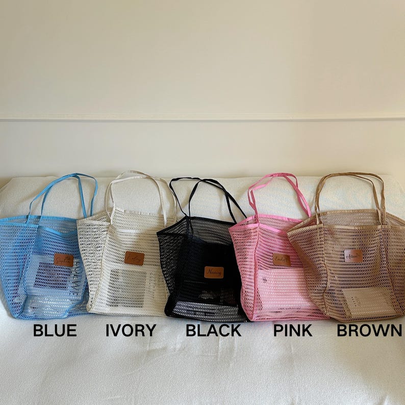 May include: Five mesh tote bags in blue, ivory, black, pink, and brown. Each bag has a rectangular leather patch and matching handles. The colors are labeled below each bag. The bags are sitting on a white surface.