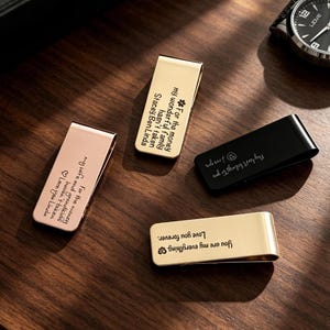 May include: Four metal money clips in gold, rose gold, and black, each engraved with a unique message. The clips are arranged on a dark wood surface, with a watch visible in the background. The messages include phrases like "Love you forever."