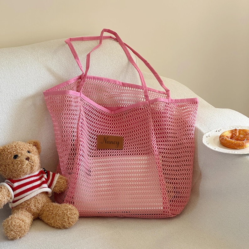 May include: A pink mesh tote bag with long handles, a rectangular shape, and a small brown leather tag that reads "Nancy". A teddy bear wearing a red and white striped shirt is next to the bag.