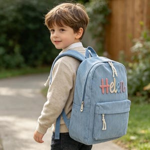 Puede incluir: Una mochila de pana azul claro con el nombre "Helen" bordado en letras coloridas. La mochila tiene un bolsillo frontal, un asa superior y correas ajustables. El ni&ntilde;o lleva un su&eacute;ter beige y vaqueros oscuros.